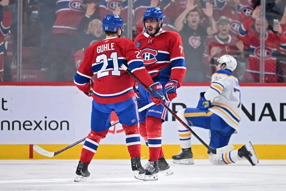 Kirby Dach yells in celebration while facing teammate Kaiden Guhle, with a Sabres player on one knee in the background.